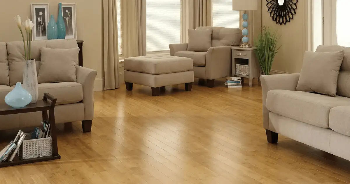 A living room with professionally installed bamboo flooring, beige sofas, a matching ottoman, a side table with a blue lamp, vases, framed pictures, and large windows with blinds and curtains in Kansas City, MO